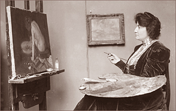 Foto av Julie Wolfthorn i sin ateljé, hon har ett staffli med en målning hos jobbar med, hon sitter med pensel i ena handen och palett i den andra och ser på målningen av en kvinna